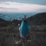 A woman with blue hair sits on a hillside, gazing at the city's twilight skyline adorned with string lights.