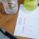 A refreshing glass of water with a green apple and workout plan on a wooden table.